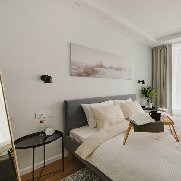 A cozy modern bedroom featuring a grey upholstered bed with soft cream bedding and layered pillows. On the bed is a wooden breakfast tray holding a book, cup, and wine glass. Minimalist black wall sconces frame a nature-themed canvas above the bed, and the space is softened with full-length curtains, potted greenery, and small round bedside tables.
