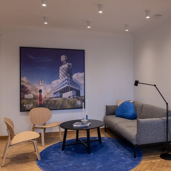 A cozy modern seating area with a gray sofa, two light wooden chairs, a black round coffee table, and a blue circular rug on wooden parquet flooring. A floor lamp provides lighting next to the sofa. A large, vibrant artwork depicting a futuristic cityscape with a child, rocket, and towering structure hangs on the wall. Minimalist decor includes a dotted blue pillow and two white candlesticks.