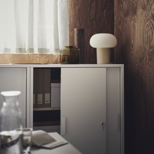 A cozy office corner with a white metal storage cabinet against a dark wood panel wall. On top of the cabinet sits a modern mushroom-shaped table lamp with a beige base and white rounded top, accompanied by two decorative glass vases—one green and one brown. Partially open cabinet doors reveal neatly organized files and storage boxes inside. A sheer white curtain filters natural light from the window above, creating a soft, ambient glow. A glass carafe and a notepad rest on a nearby desk in the foreground, completing the tranquil and organized workspace.