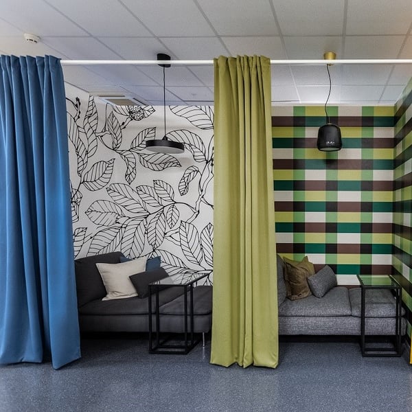 A cozy office lounge area is divided into two semi-private nooks using ceiling-mounted curtains—one blue and the other green. Each nook contains a gray sofa with assorted throw pillows and a black side table. The left nook features a white wall with a black leaf pattern, while the right has a bold, checkered pattern of green, brown, yellow, and teal stripes. Black pendant lights hang over each seating area, and the space has a contemporary and playful vibe, ideal for casual meetings or relaxation.