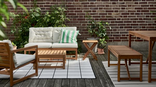 A cozy outdoor living area with wooden furniture, including a cushioned sofa, armchair, coffee table, side table, and a dining bench set – arranged on a stylish rug with green plants adding a touch of nature.