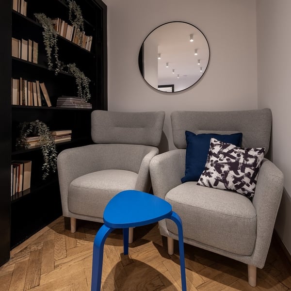 A cozy reading nook featuring two light gray armchairs with high backs and wooden legs, accented by two decorative pillows in navy and black-and-white patterns. A small bright blue triangular side table stands between the chairs. Behind them is a tall black bookshelf filled with books and draped greenery. A round wall mirror reflects ceiling lights from the opposite room. The floor is parquet wood.