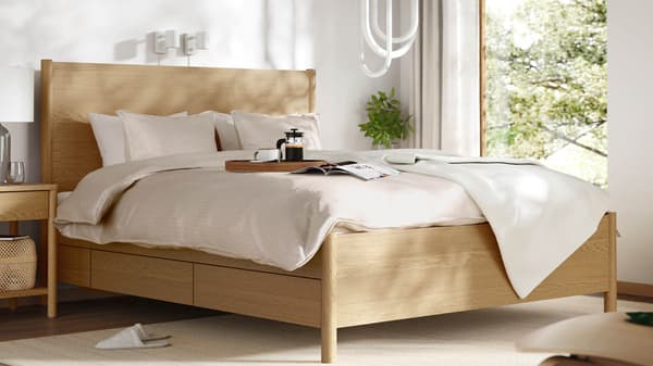 A cozy, sunlit bedroom featuring a wooden bed with beige bedding, a bedside table, and a tray with coffee and a magazine on the bed.