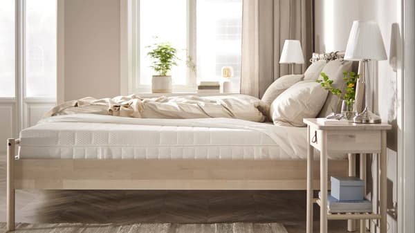 A cozy, sunlit bedroom with a neatly made bed, beige bedding, a wooden nightstand, and soft natural light from large windows.