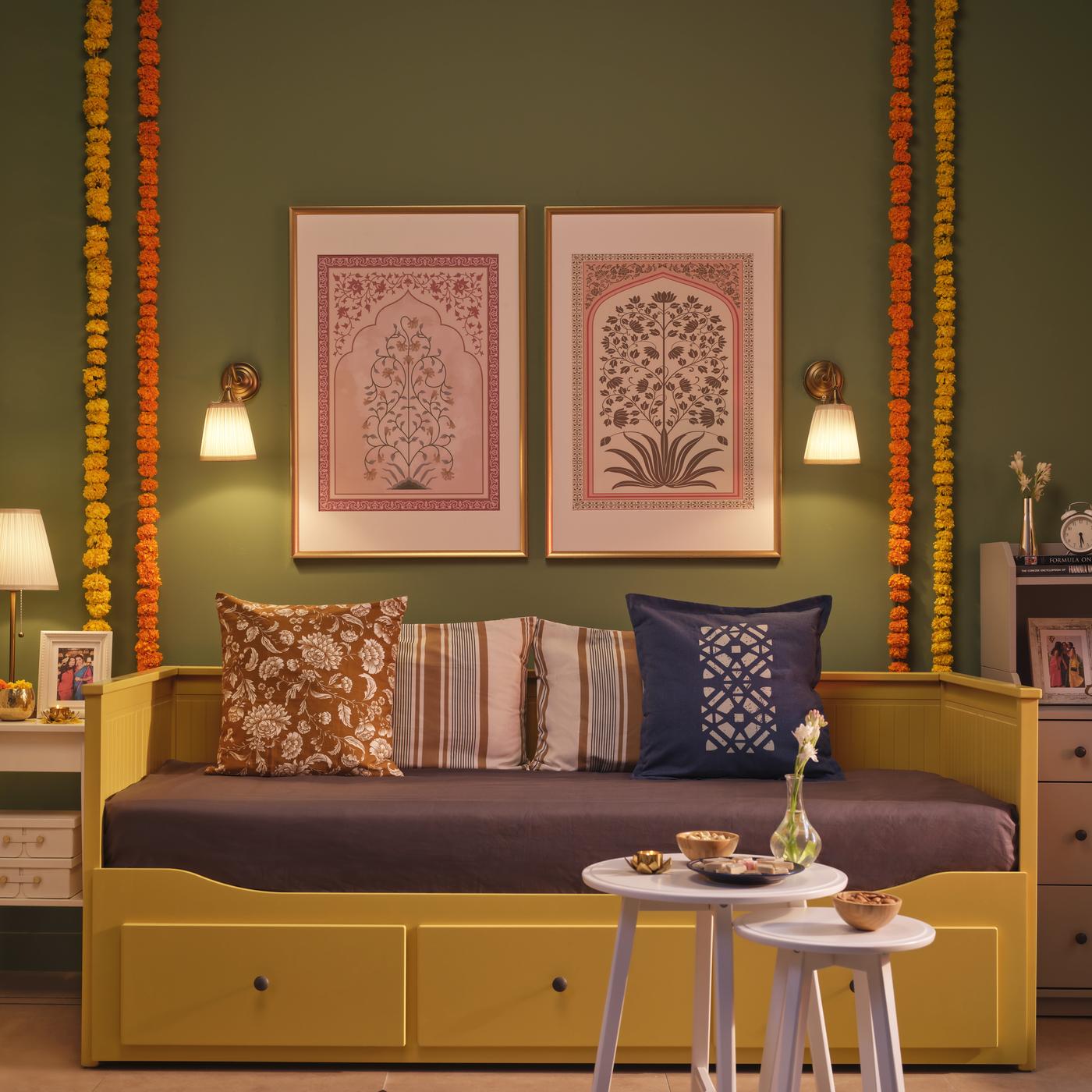 A cozy, well-decorated living space featuring a yellow HEMNES daybed with cushions of assorted patterned throw pillows in earthy tones. Above the bed are two framed botanical prints, flanked by wall lamps and strings of marigold flowers.