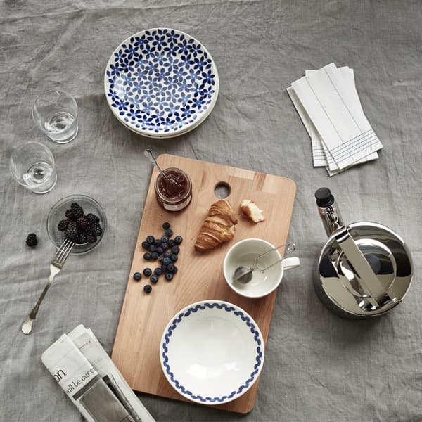 A croissant, jam and berries on a wooden board with patterned plates and bowls, a teapot and a newspaper.