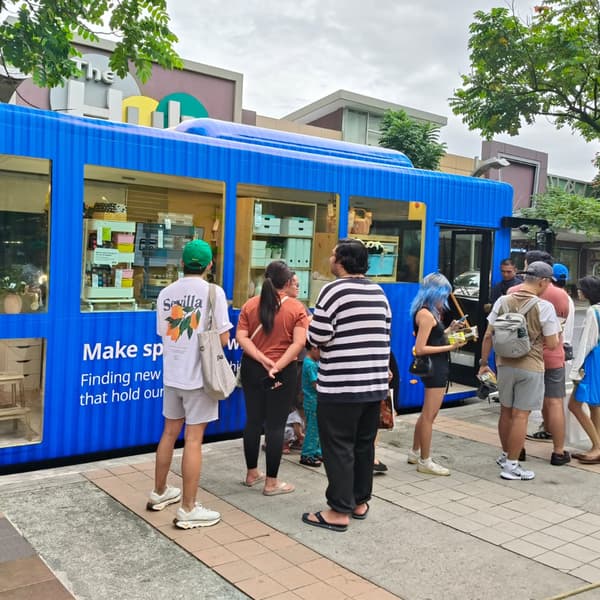 A crowd lines up at a vibrant blue mobile store, showcasing products, with trees and buildings in the background.