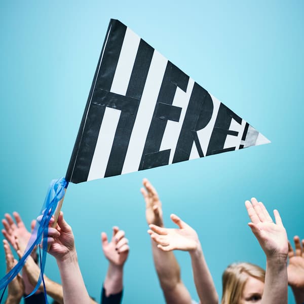 A crowd of people have their hands in the air, while one person waves a handmade flag that says ‘here.’
