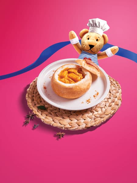 A curry chicken in bread bowl with a FABLER BJÖRN teddy bear soft toy wearing a white chef hat while raising its up in the air on a pink background.