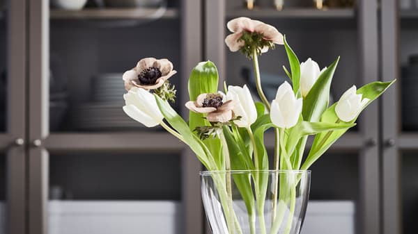 A curvy VASEN vase with a small bouquet of white tulips, brownish poppies and fresh green leaves in front of a cupboard.