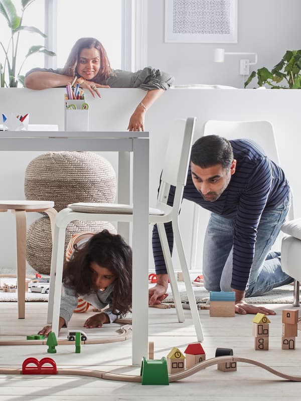 A dad down on his knees playing with his daughter under the dining room table while mum looks on from the sofa.