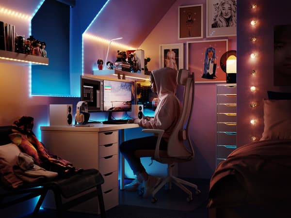 A dark bedroom workspace area lit with blue and string lights, a child sits at a gaming desk playing.