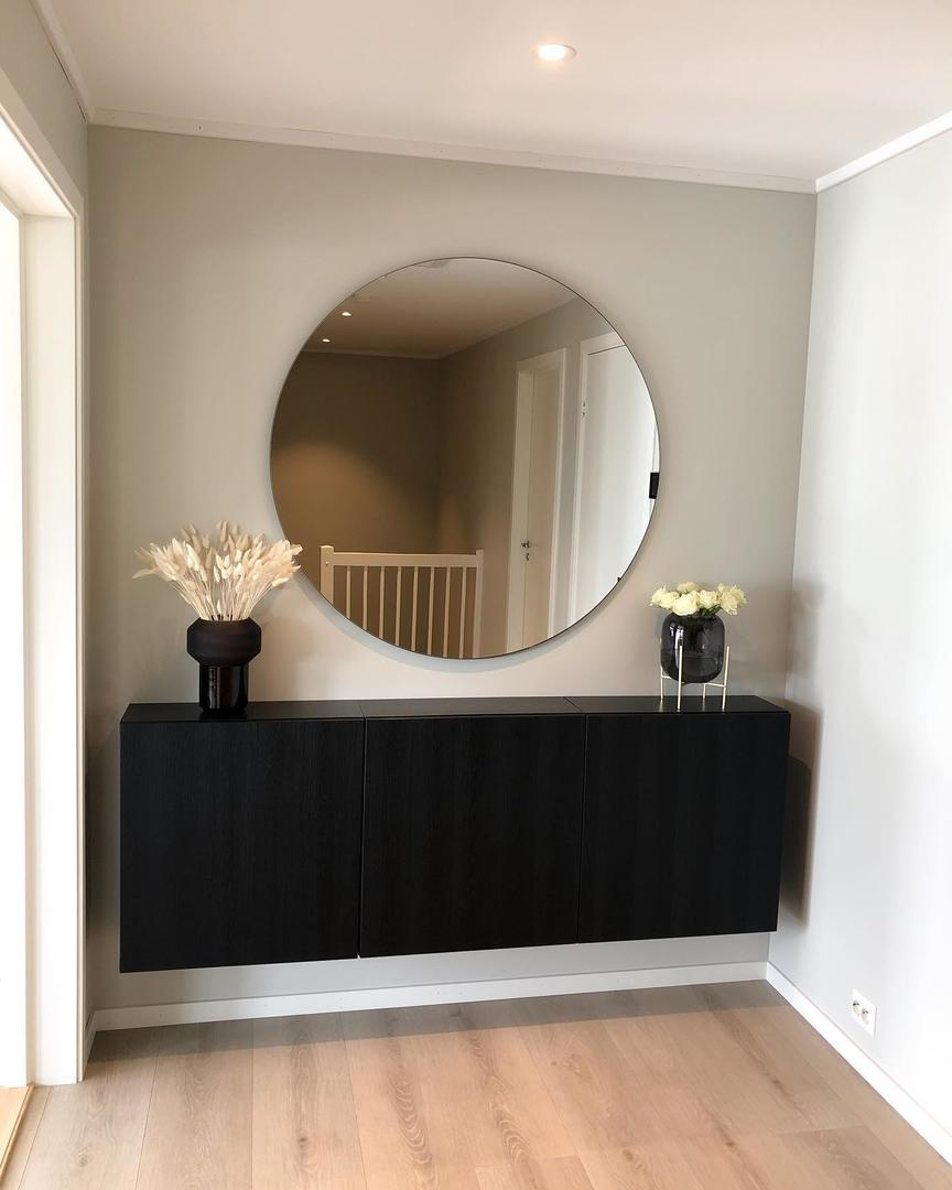 A dark BESTÅ storage unit in a hallway, entryway or landing with a large round mirror above it.