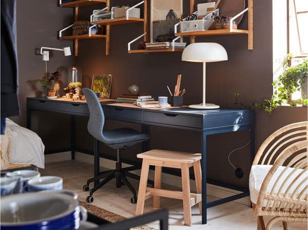 A dark blue desk, a wall-mounted shelf combination in bamboo, a white table lamp and a birch stool.