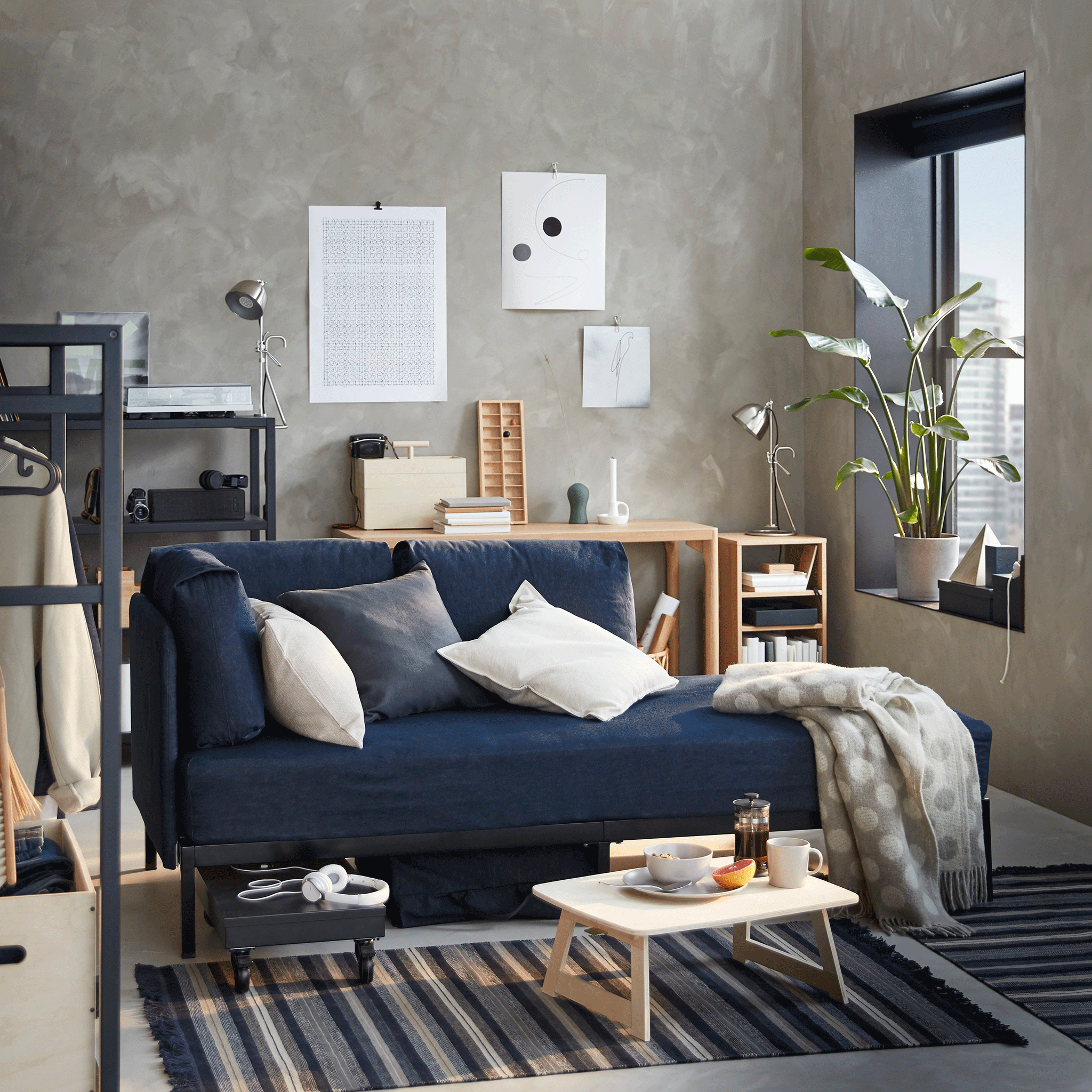 A dark blue RÅVAROR daybed is set against a concrete-style wall. A RÅVAROR storage bag has been hung above it.