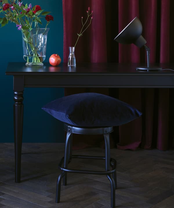 A dark blue velvet cushion perched on a stool, in front of a black wood desk and dark red curtain.