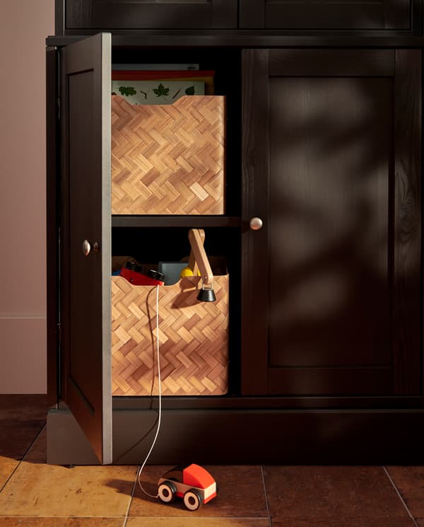 A dark brown base cabinet has one door open, inside are two shelves with two bamboo boxes with books and children’s toys.