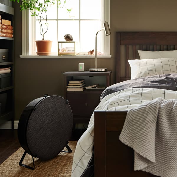 A dark brown IDANÄS bed with checked VITKLÖVER bed linen on a natural LOHALS flatwoven rug, plus a round black air purifier.