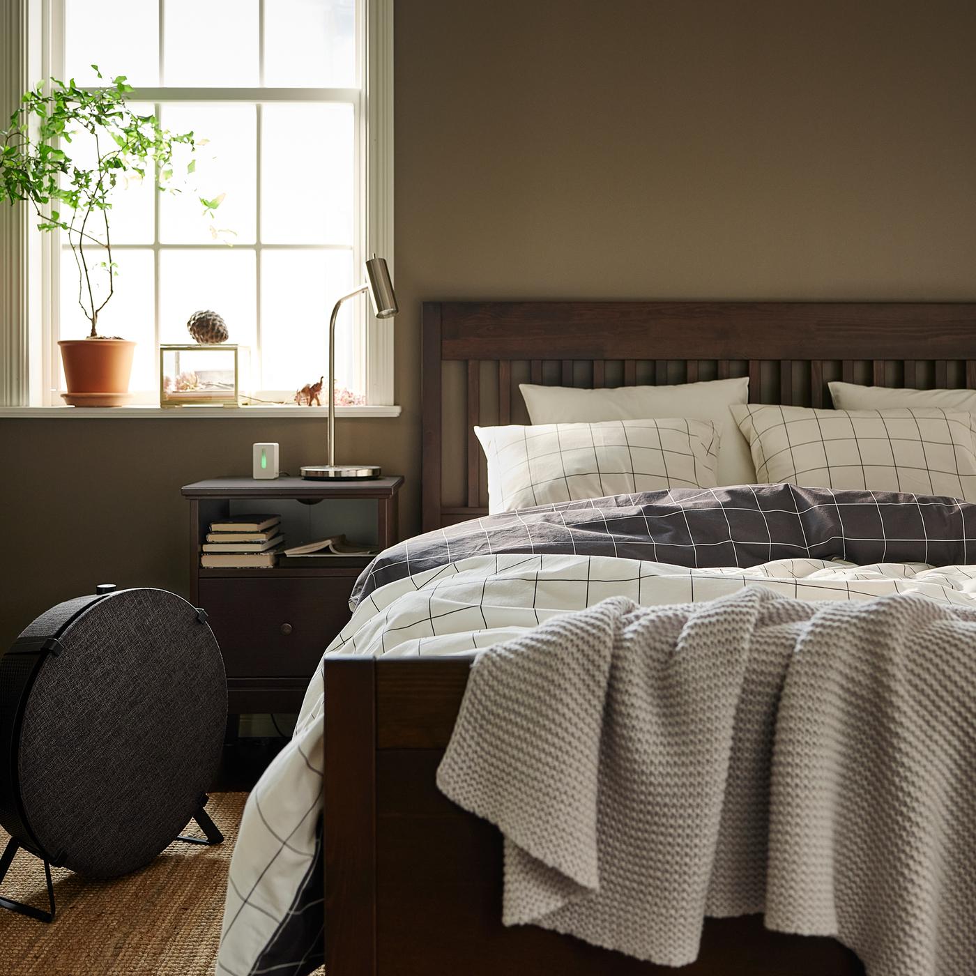 A dark brown IDANÄS bed with VITKLÖVER bed linen stands on a flatwoven LOHALS rug near a black STARKVIND air purifier.