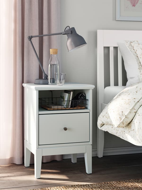 A dark desk lamp standing on a white IDANÄS bedside table next to a white IDANÄS double bed in a dimly sunlit, light bedroom.