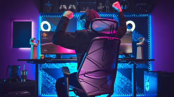 A dark gaming room with a lit-up gaming station. A person, sitting in a gaming chair, is raising the arms victoriously. 