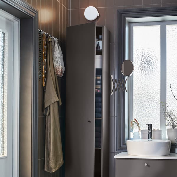 A dark gray high cabinet, a round mirror with an extendable arm, knobs in chrome effect and a dark gray wash-stand.