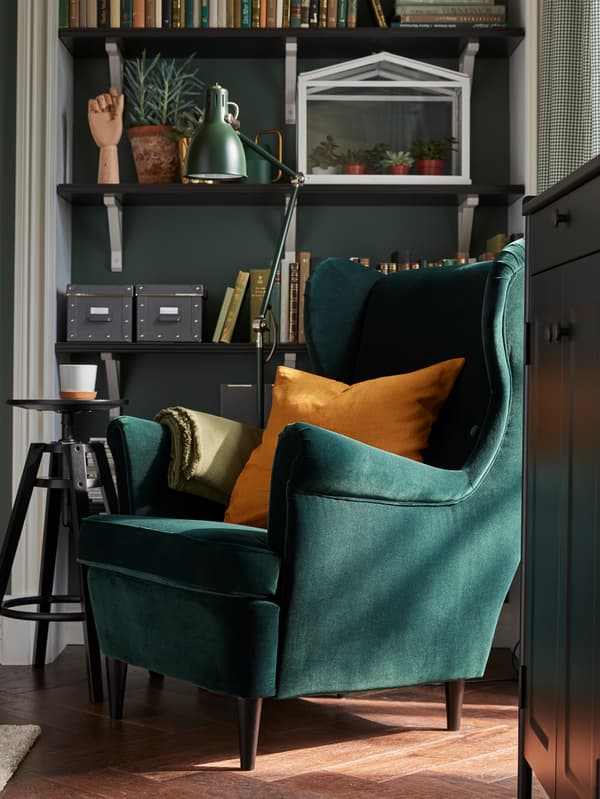 A dark green STRANDMON armchair with an ochre cushion and an angle-poised lamp and shelves with books and plants.