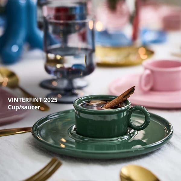 A dark green VINTERFINT cup with saucer with a VINTERSAGA mulled fruit drink and cinnamon stick in it is on a festive table.