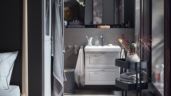 A dark grey bathroom with a wash-stand in light grey, a black RÅSKOG trolley with folded towels and decorative flowers.