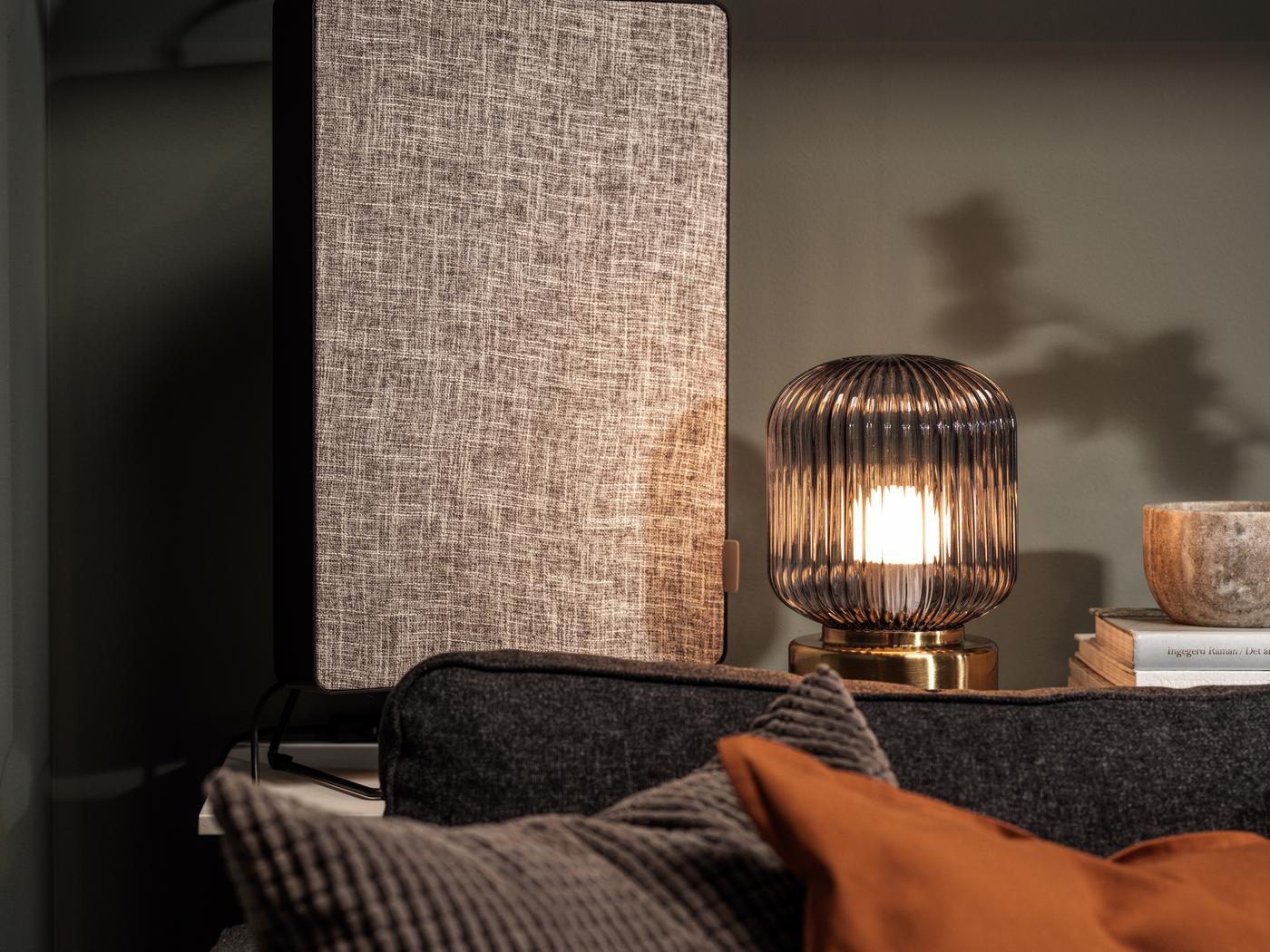 A dark grey FÖRNUFTIG air purifier stands on a small table next to a glass lamp, behind a brown sofa with pillows.