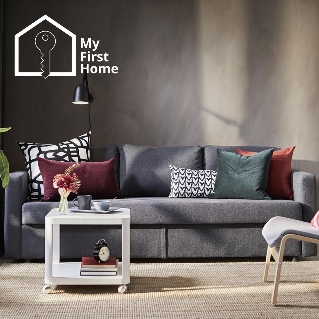 A dark grey FRIHETEN 3-seat sofa is placed on top of natural flatwoven rug in the living room, in front of a white TINGBY side table on castors. The top left corner shows the "My First Home" icon, which consists of two parts: an outlined key placed on the outlined silhouette of a house, and the text "My First Home" placed on the right of the graphics.