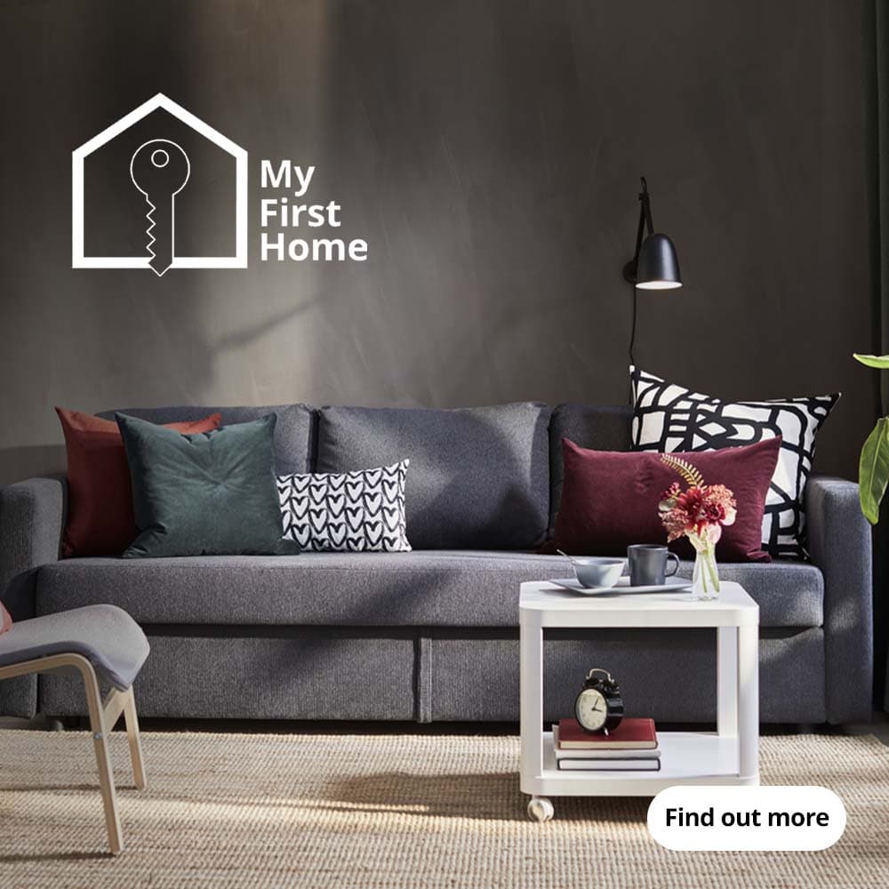 A dark grey FRIHETEN 3-seat sofa is placed on top of natural flatwoven rug in the living room, in front of a white TINGBY side table on castors. The top left corner shows the "My First Home" icon, which consists of two parts: an outlined key placed on the outlined silhouette of a house, and the text "My First Home" placed on the right of the graphics.