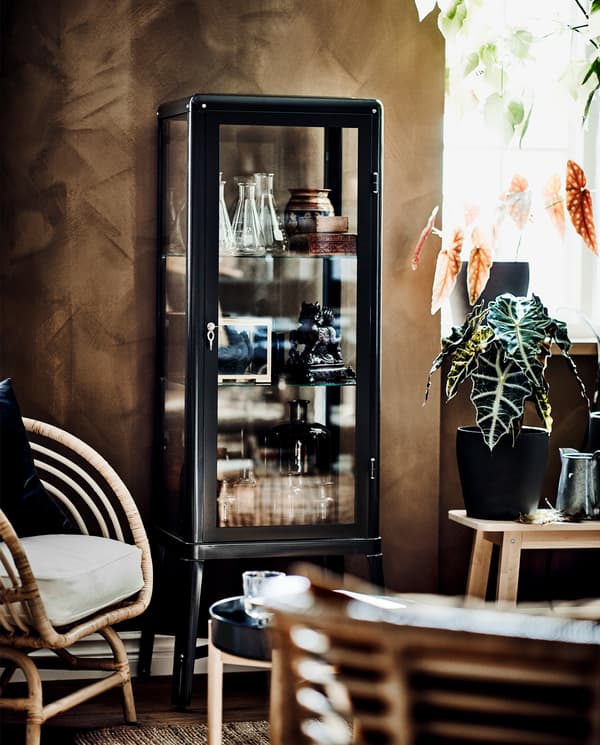 A dark grey glass-door cabinet with decorative items stored inside, a green plant and a rattan armchair with a white cushion.