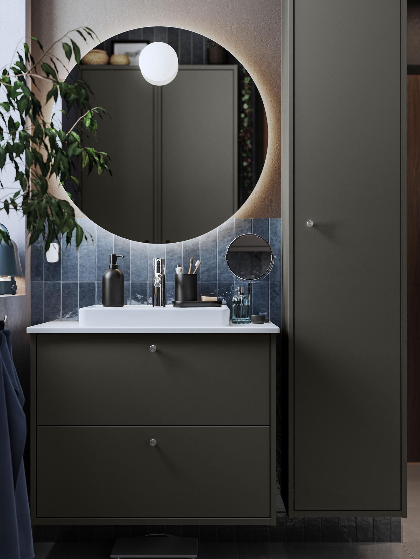 A dark grey HAVBÄCK washstand with drawer and a white ORRSJÖN semi-recessed wash-basin are placed in a dark-tiled bathroom.