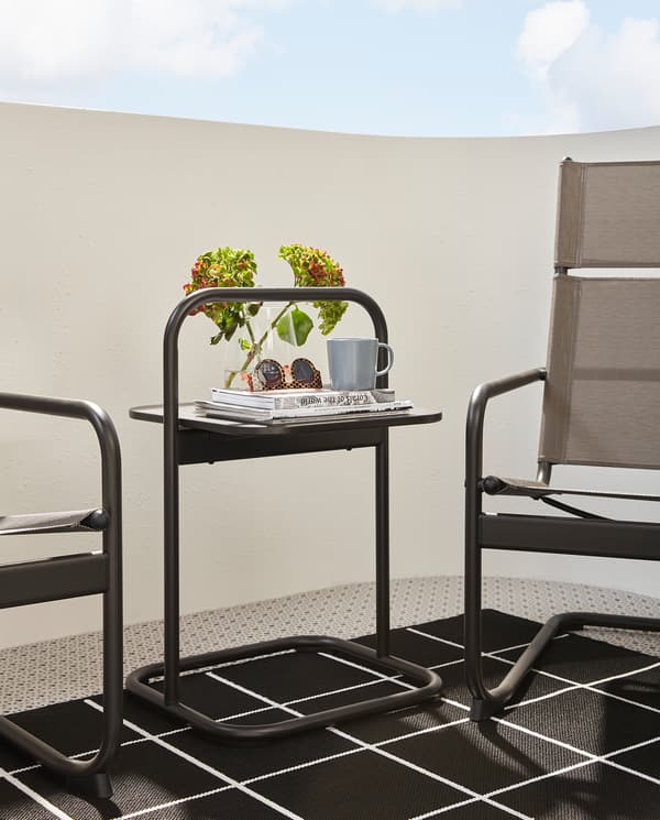 A dark grey HUSARÖ side table with handle stands on a rug between two armchairs. Sunglasses and a mug stand on the tabletop.