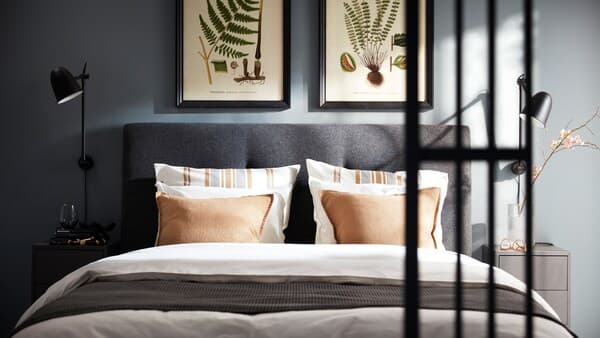 A dark grey IDANÄS bed with white ÄNGSLILJA bed linen, a dark grey VÅRELD bedspread, and black SKURUP wall lamps on each side.