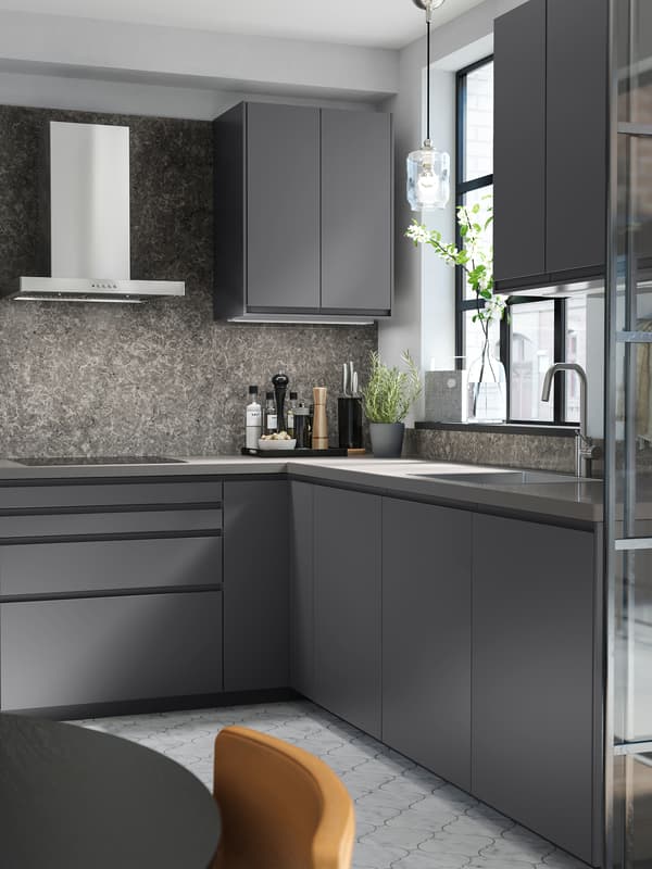 A dark grey kitchen with dark grey cupboards, a stainless steel wall-mounted extractor hood and patterned flooring.