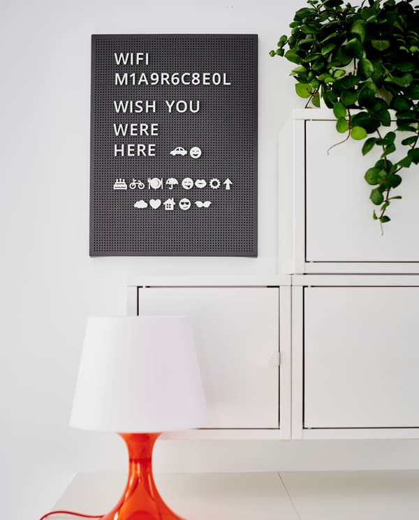 A dark grey pegboard with letters and symbols, a white/orange table lamp, three wall-mounted cabinets and a green plant.