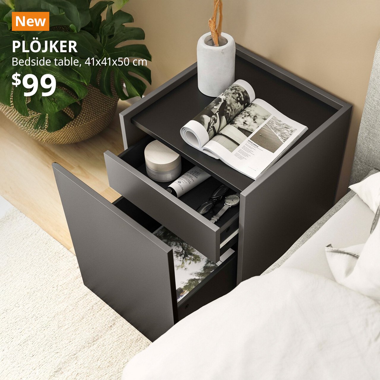 A dark grey PLÖJKER bedside table placed next to a bed in a bedroom setting. An orange "NEW" label is on the top left of the image with black text that reads: PLÖJKER, Bedside table, 41x41x50 cm, Price is $99".