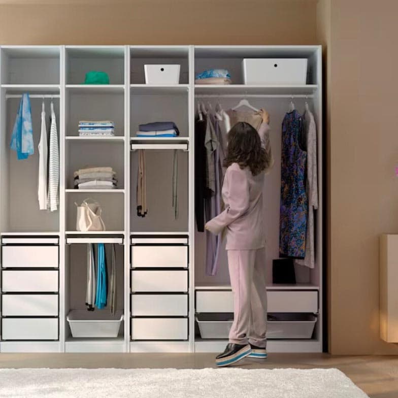A dark-haired woman hangs a dress on the clothes rail of a white, open PAX wardrobe combination in a room with brown walls.