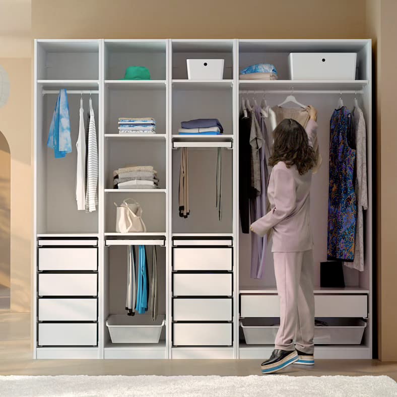 A dark-haired woman in a pink trouser suit hangs a dress on the clothes rail of a white, open PAX wardrobe combination.