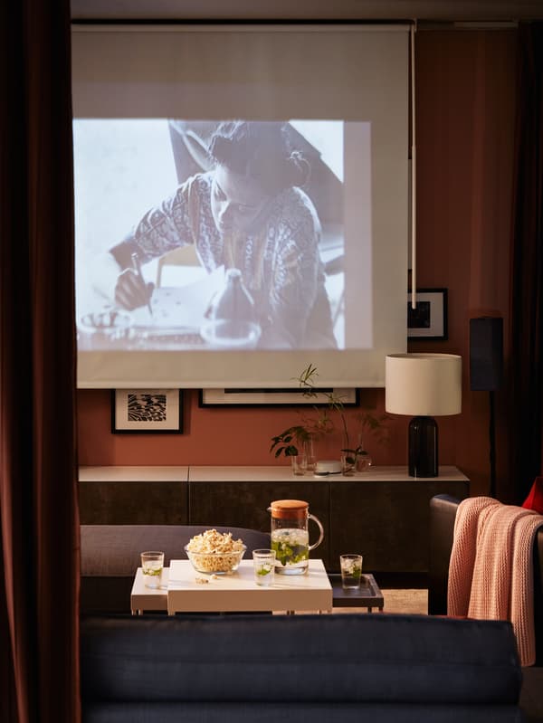 A dark living room with a movie projected on a block-out roller blind, a TV bench with doors, popcorn.