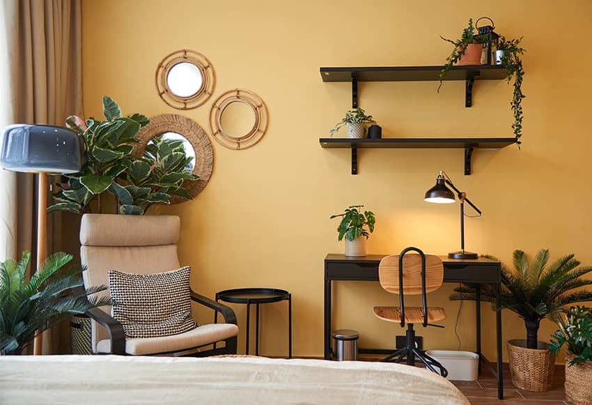 A dark yellow room showcasing a small desk with a lamp shining on it, a light brich armchair and white bed.