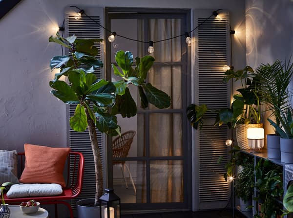 A darkened balcony space with plants, lit with an outdoor table lamp, a lantern and a SVARTRÅ LED black lighting chain.