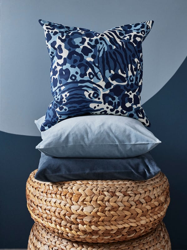 A deep blue KAPHYACINT cushion with print patterns is propped on cushions in light blue and dark blue. The cushions are placed on a handwoven basket, against a blue background.