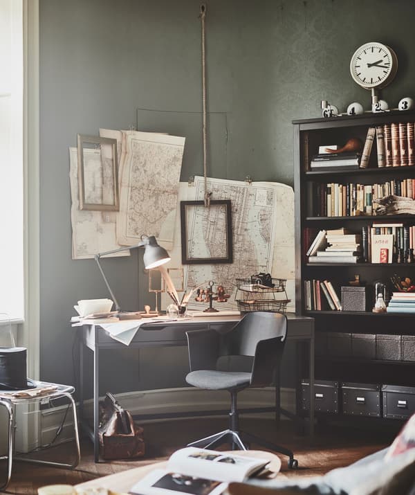 A desk and chair in the corner of the living room, with maps and frames on the wall and an IKEA HEMNES bookcase.