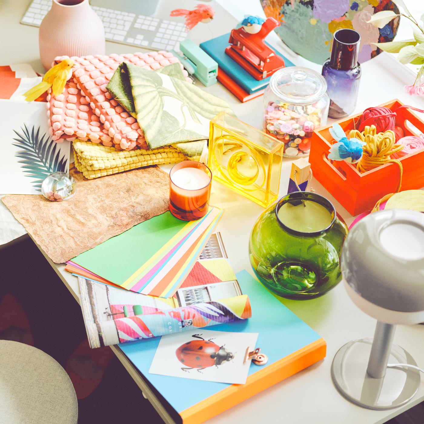 A desk holds a messy mix of colourful accessories, vases, cushion covers and a beige BLÅSVERK table lamp.
