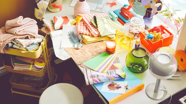 A desk is covered with colorful cushion covers, paper and a green vase. A yellow RÅSKOG cart is to the side.
