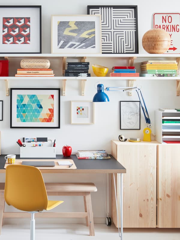 A desk on castors is placed next to a wall, surrounded by light wood IVAR cabinets, wall shelves and framed artwork.