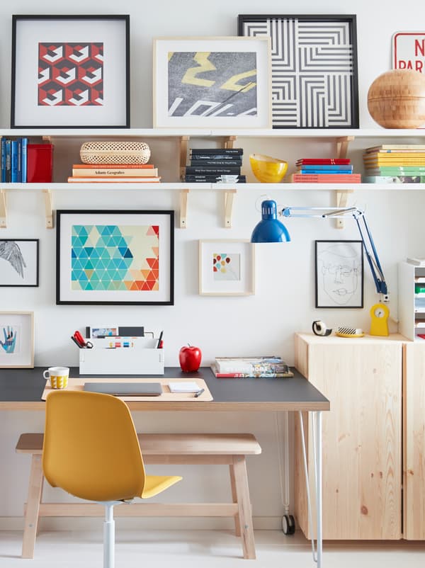A desk on castors stands by a wall, surrounded by a TERTIAL work lamp, wooden cabinets, wall shelves, and framed artwork.